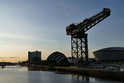 Cranes at riverbank against sky during sunset