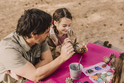 Smiling girl coloring pine cone with father while sitting at picnic table in park