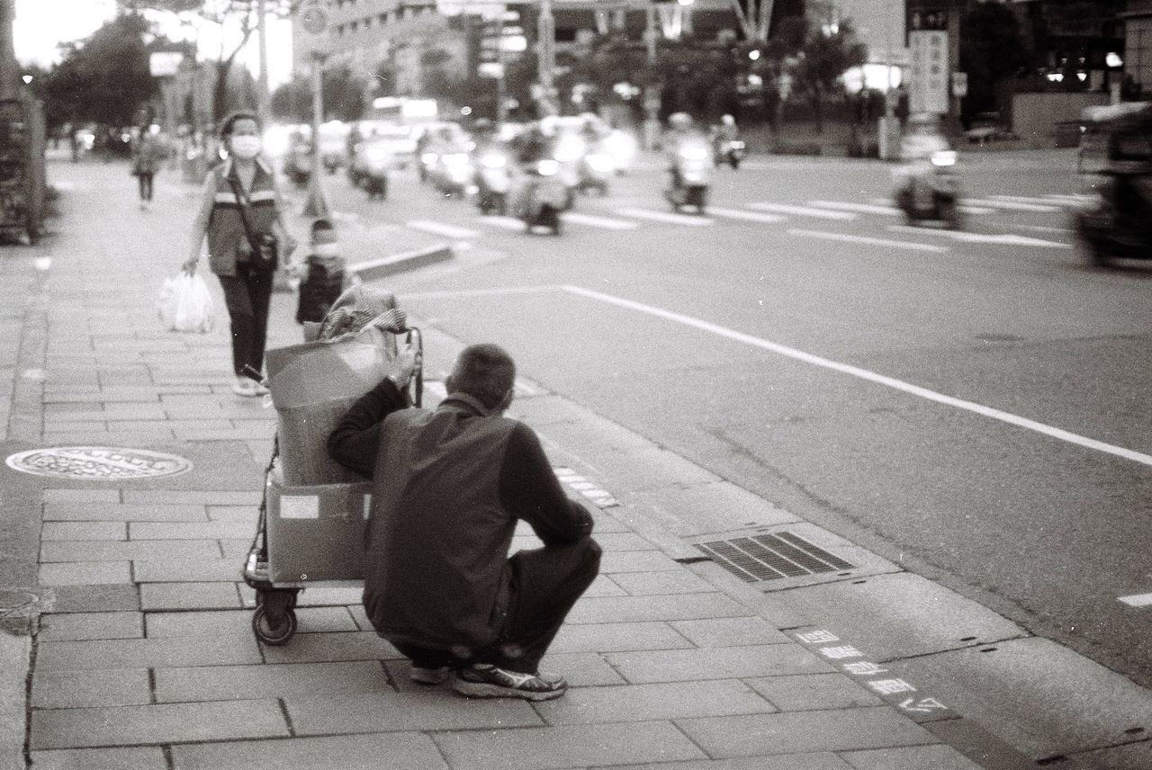 REAR VIEW OF MAN SITTING ON SIDEWALK BY STREET