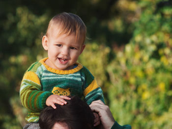 Portrait of cute boy sitting on father neck against tree