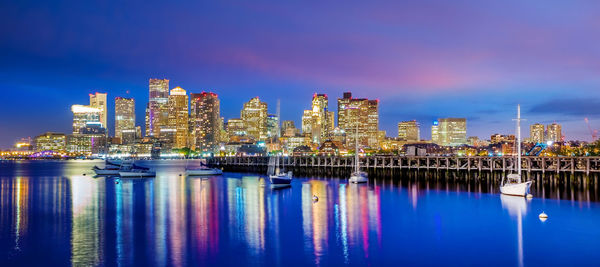 Illuminated buildings by river against blue sky at night