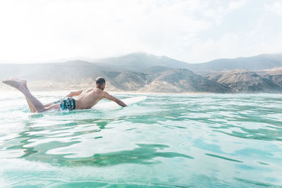 Full length of shirtless man surfing in sea against mountains