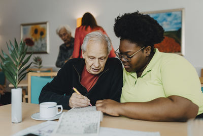 Nurse sitting next to senior man solving sudoku puzzle at retirement home
