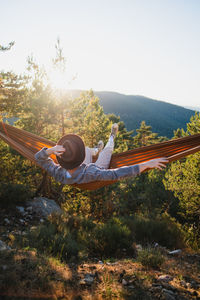 Back view of unrecognizable male in hat lying in hammock while resting in forest with green trees on sunny summer day