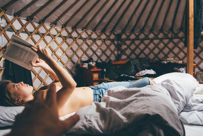 Cropped image of person hand reaching towards topless woman reading book on bed in tend