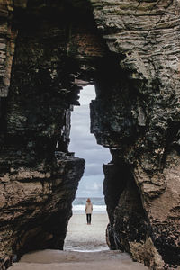 View of distant anonymous traveler in outerwear through massive stony formation on sandy coast near sea in nature of spain