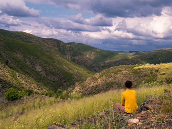 Rear view of woman sitting on mountain against cloudy sky