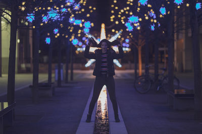 Portrait of woman standing on footpath against illuminated lights at night