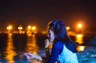 Side view of woman smoking cigarette while standing against river at night