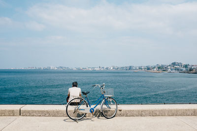 Rear view of man with bicycle on promenade by sea against cloudy sky