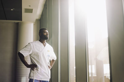Low angle view of male medical professional standing with arms akimbo near window in hospital