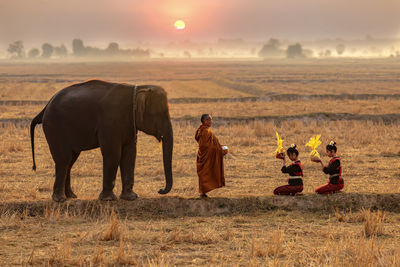 Full length of monk with people and elephant on field during sunrise