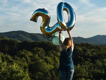 Rear view of woman with helium balloons standing on mountain against sky