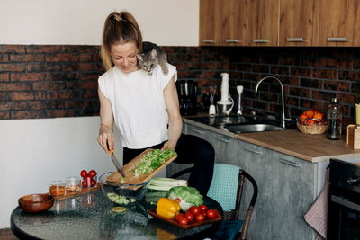 Young woman preparing vegetarian green salad at home and playing with cat