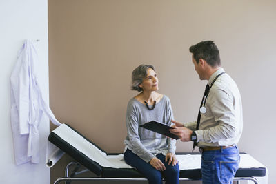 Senior patient talking with doctor while sitting on bed against wall at hospital