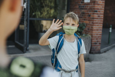 Little boy wearing protective face mask waving his brother outside school building