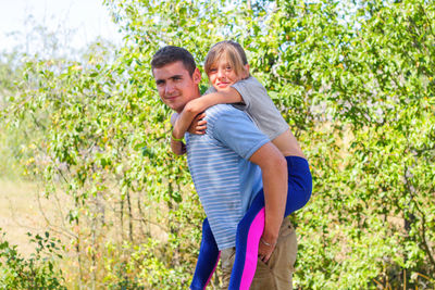 Brother giving sister ride on back in forest. portrait of happy girl on young man shoulders, piggy