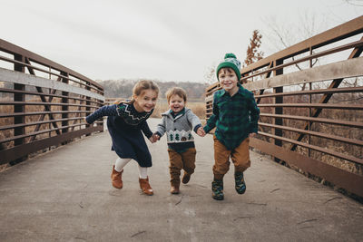 Three siblings holding hands running on bridge toward camera
