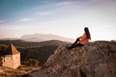 Woman sitting on rock looking at mountain against sky