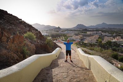 Portrait of man with arms outstretched standing on footpath against sky