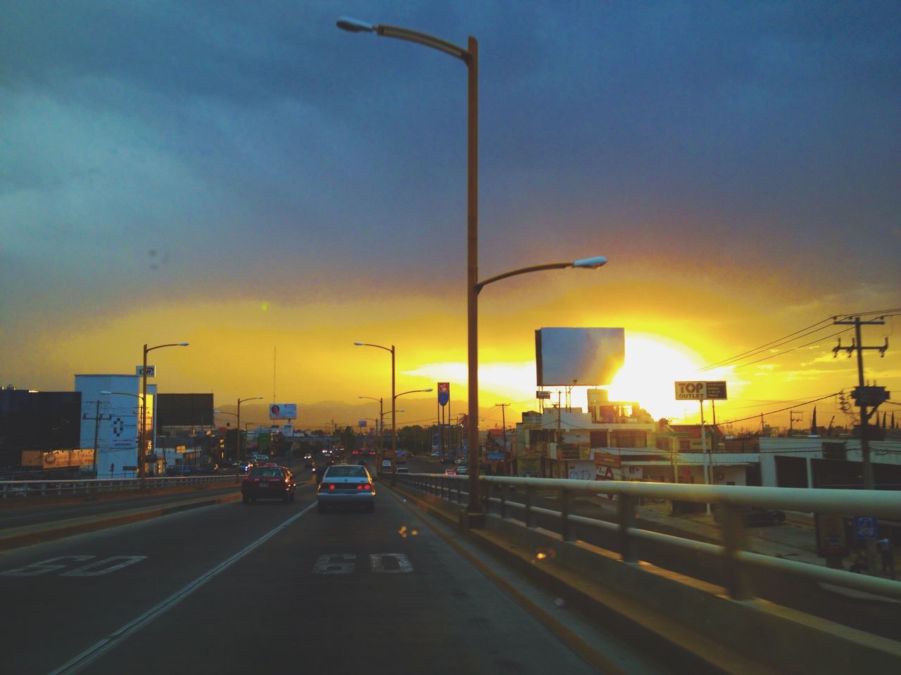 transportation, sunset, car, street light, road, sky, mode of transport, land vehicle, orange color, built structure, street, architecture, illuminated, the way forward, cloud - sky, road marking, lighting equipment, building exterior, outdoors, city