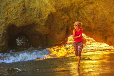 Full length of mature woman wearing sunglasses running in sea against mountain during sunset