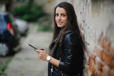 Young woman is smiling and holding phone by the brickwall.