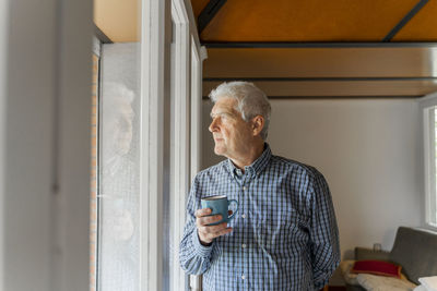 Serious senior man looking at home through window