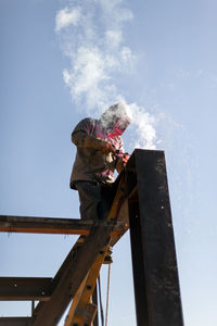 Welder arc-welds structural steel i-beam intersection from a ladder