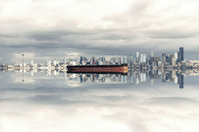 Modern buildings reflecting on river against cloudy sky