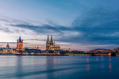 Illuminated city at waterfront against cloudy sky