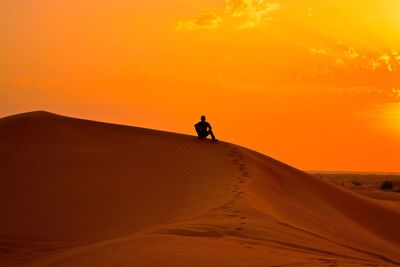 Silhouette man sitting sand dune in desert against orange sky