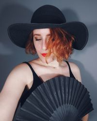 Beautiful woman holding hand fan and wearing hat against gray background