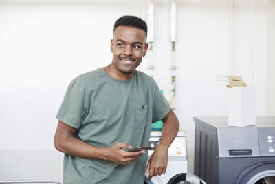 Smiling man holding mobile phone while standing at laundromat