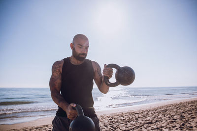 Man exercising with kettlebells at beach against sky