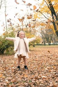 Happy little child girl in coat and dress throwing yellow leaves up in the air in autumn park