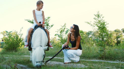 A woman and a boy are walking around the field, son is riding a pony, mother is holding a pony for