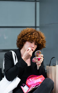 A non-binary person applies makeup sitting on a bench