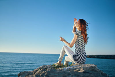 Woman sitting on rock by sea against clear blue sky