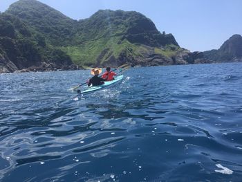 Rear view of people kayaking over sea by mountains against clear sky