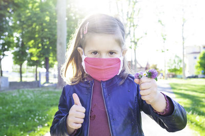 Cute little girl shows thumb up, wearing protective mask and holding bunch of wildflowers 
