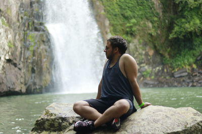 Young man sitting on rock against waterfall
