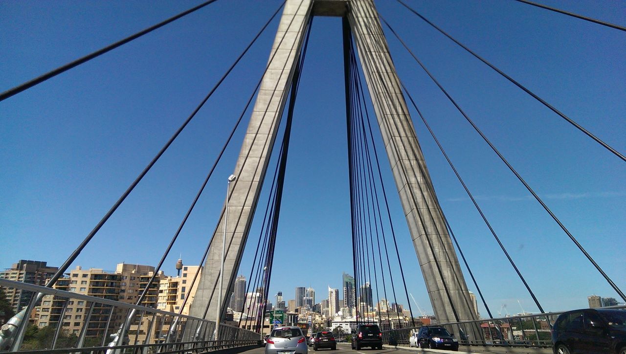 transportation, built structure, architecture, blue, connection, low angle view, suspension bridge, city, building exterior, engineering, bridge - man made structure, clear sky, mode of transport, cable, car, sky, cable-stayed bridge, modern, outdoors, travel