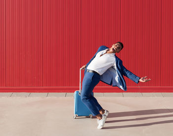 Full length side view of happy young african american male entrepreneur in stylish outfit with suitcase screaming and jumping near red wall while celebrating business success