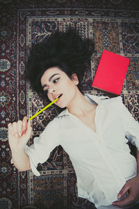 High angle view of woman lying with pencil in mouth by red book on carpet