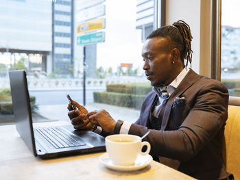 Side view of concentrated african american businessman in suit sitting at table with coffee in cafe and browsing smartphone while working on remote project