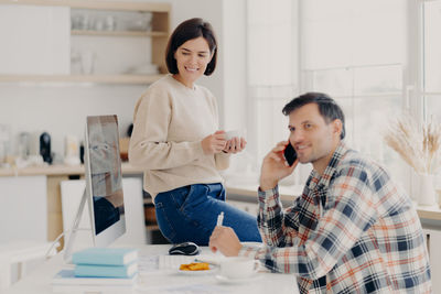 Smiling woman sitting on table while man talking over smart phone at home