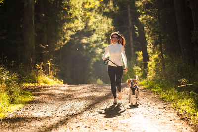 Full length of woman running with dog on road at forest