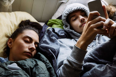 High angle view of young man using mobile phone while lying by friend on bed at home