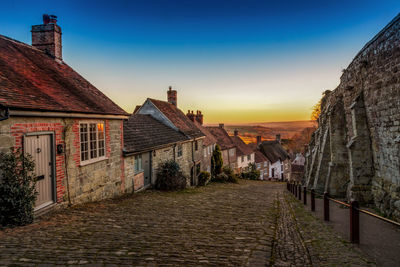 Footpath amidst houses and buildings against sky at sunset